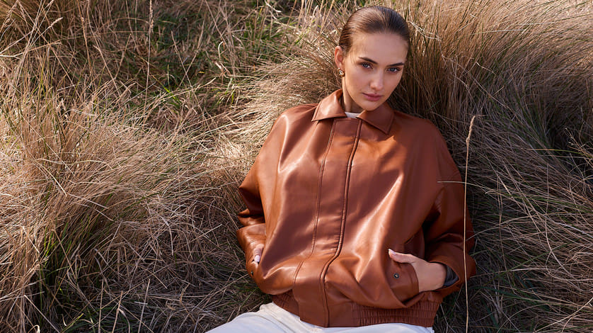 Model in a cognac leather bomber jacket white top and trousers lying in sunlit golden tall grass looking at the camera