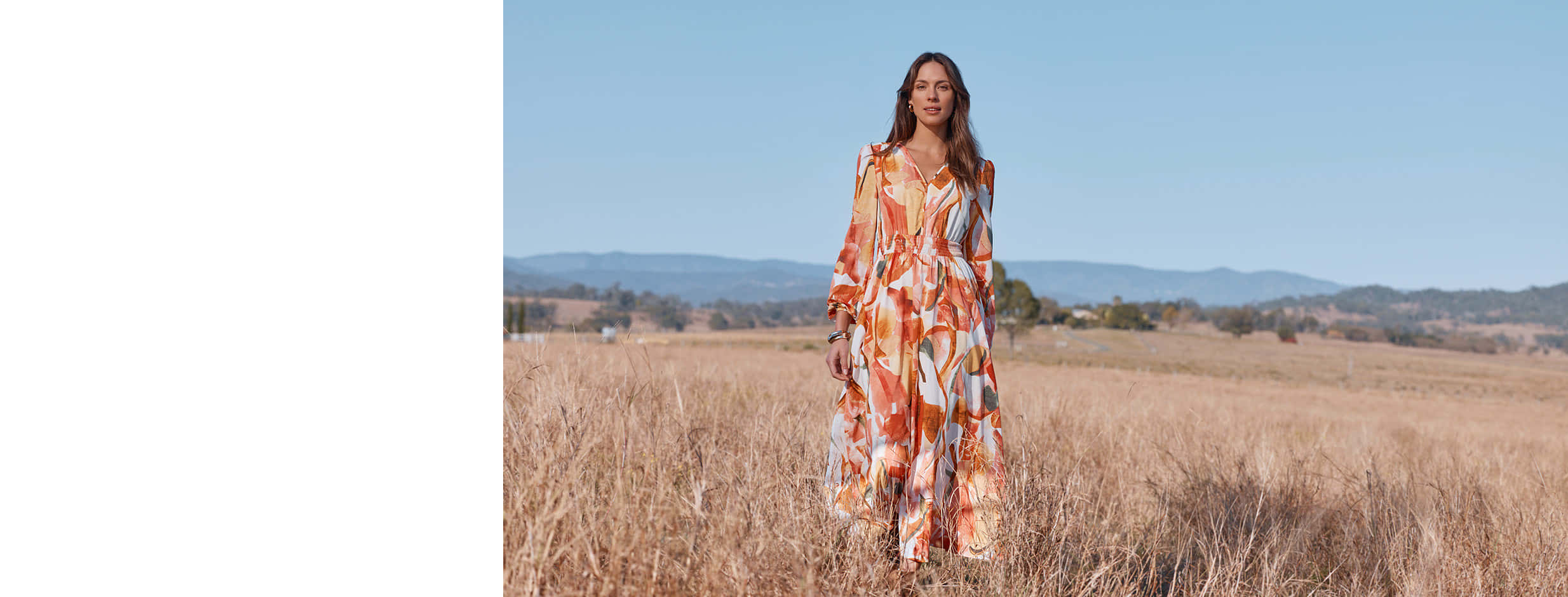 Model in an orange abstract-print long chiffon dress standing in a golden meadow with mountains in the distance