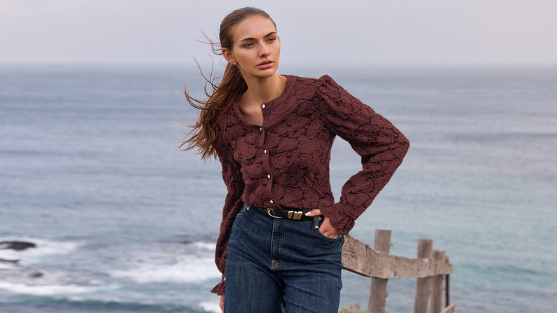 Model in a burgundy floral puff-sleeve blouse high-waisted blue jeans and brown belt standing by a wooden fence by the sea