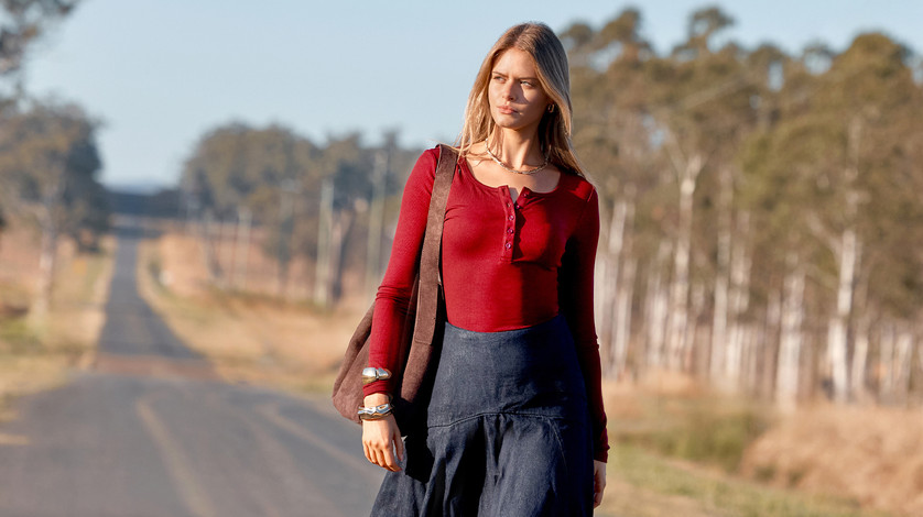 Blonde model in a dark-red henley long-sleeve top and navy long skirt with a brown shoulder bag walking on a country road