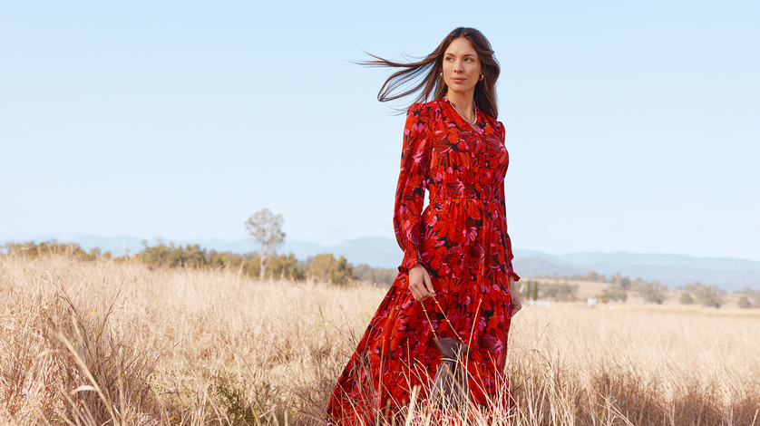 Dark-haired model in a red printed long-sleeve chiffon maxi dress standing in a golden open field with wind-blown hair and blue sky