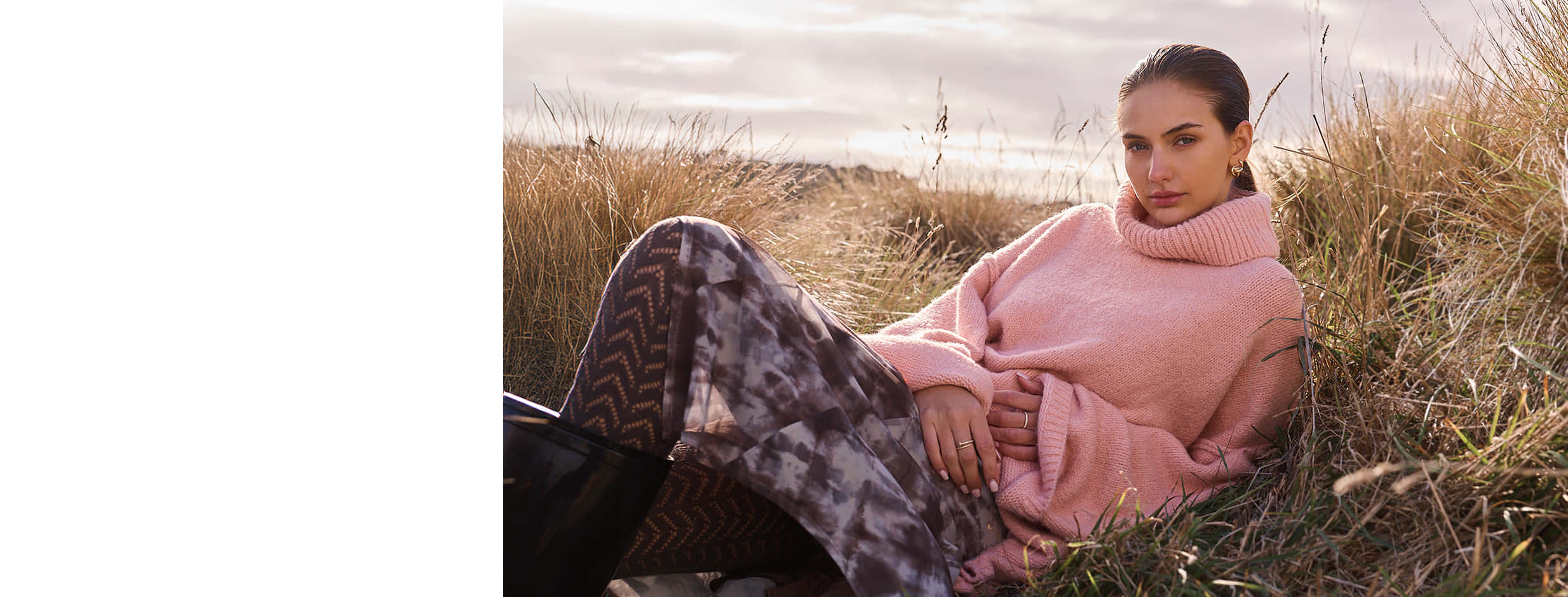 Model in a chunky pink turtleneck sweater and printed brown pants reclining in golden coastal grass under a soft overcast sky
