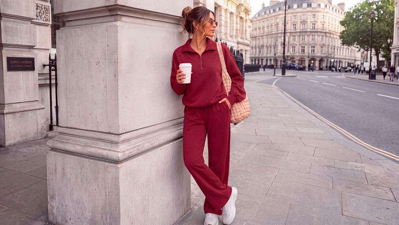 Model in a burgundy half-zip sweatshirt and matching sweatpants with white sneakers holding a coffee leaning on a city street building