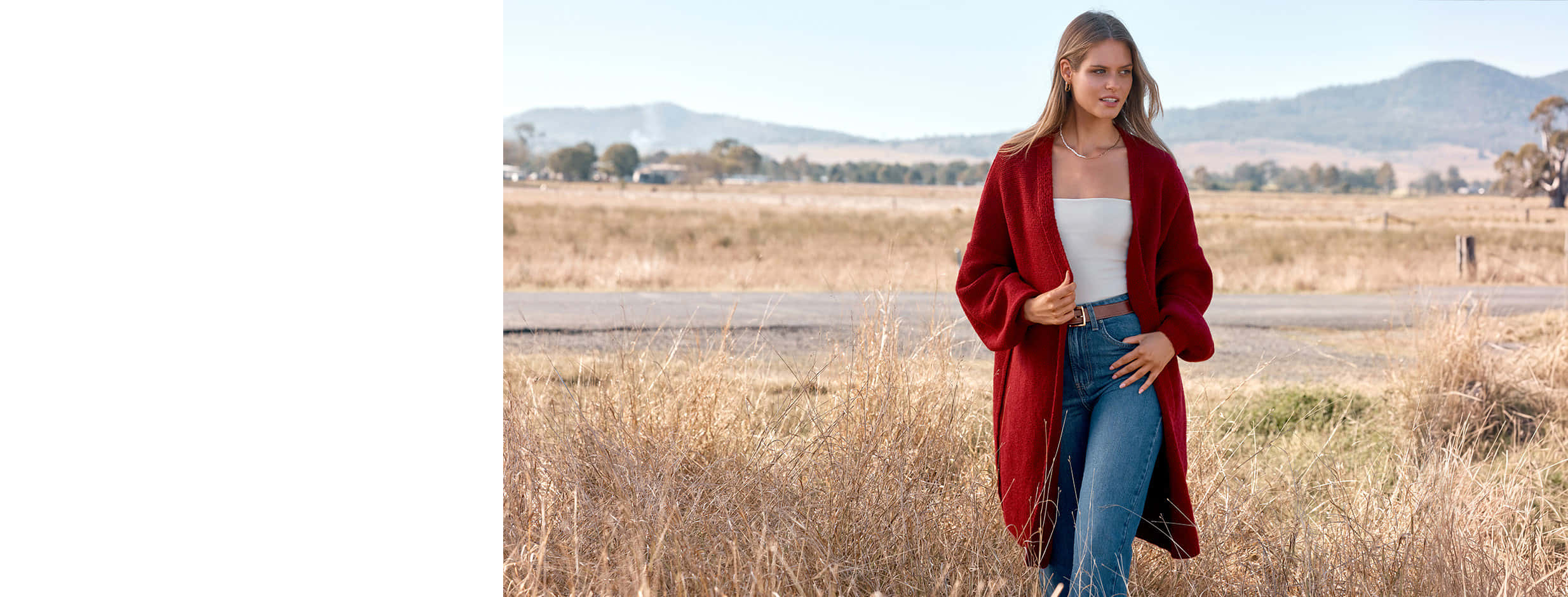 Model in a bright-red long knit cardigan white top and blue jeans walking through golden autumn grassland with hills behind